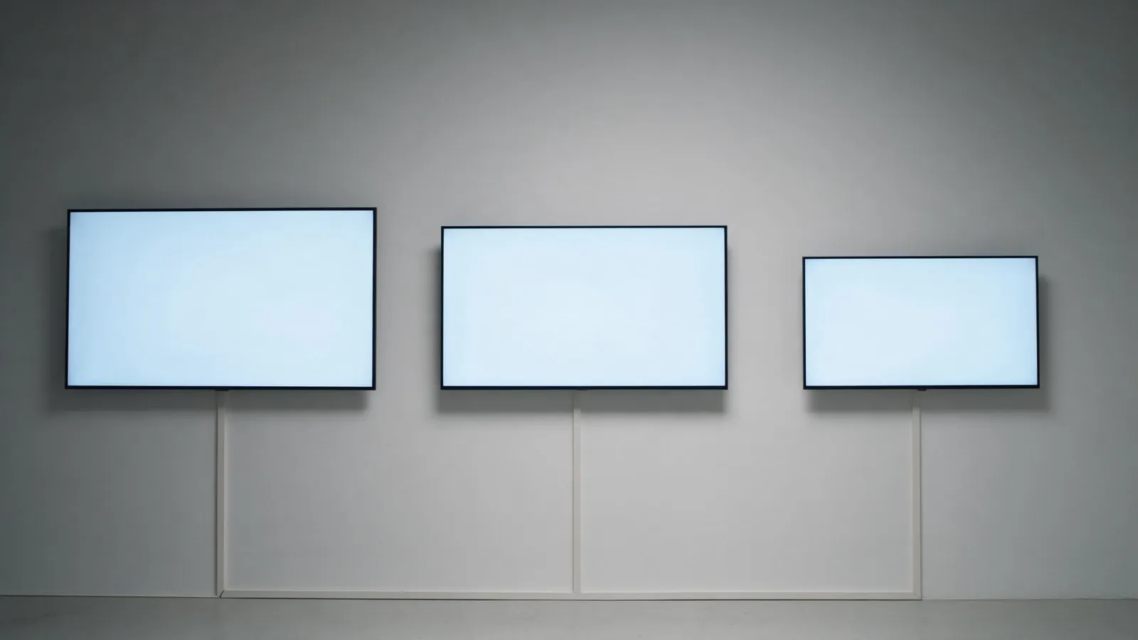 Three wall mounted flat panel displays of clearly different sizes side by side on a plain studio wall